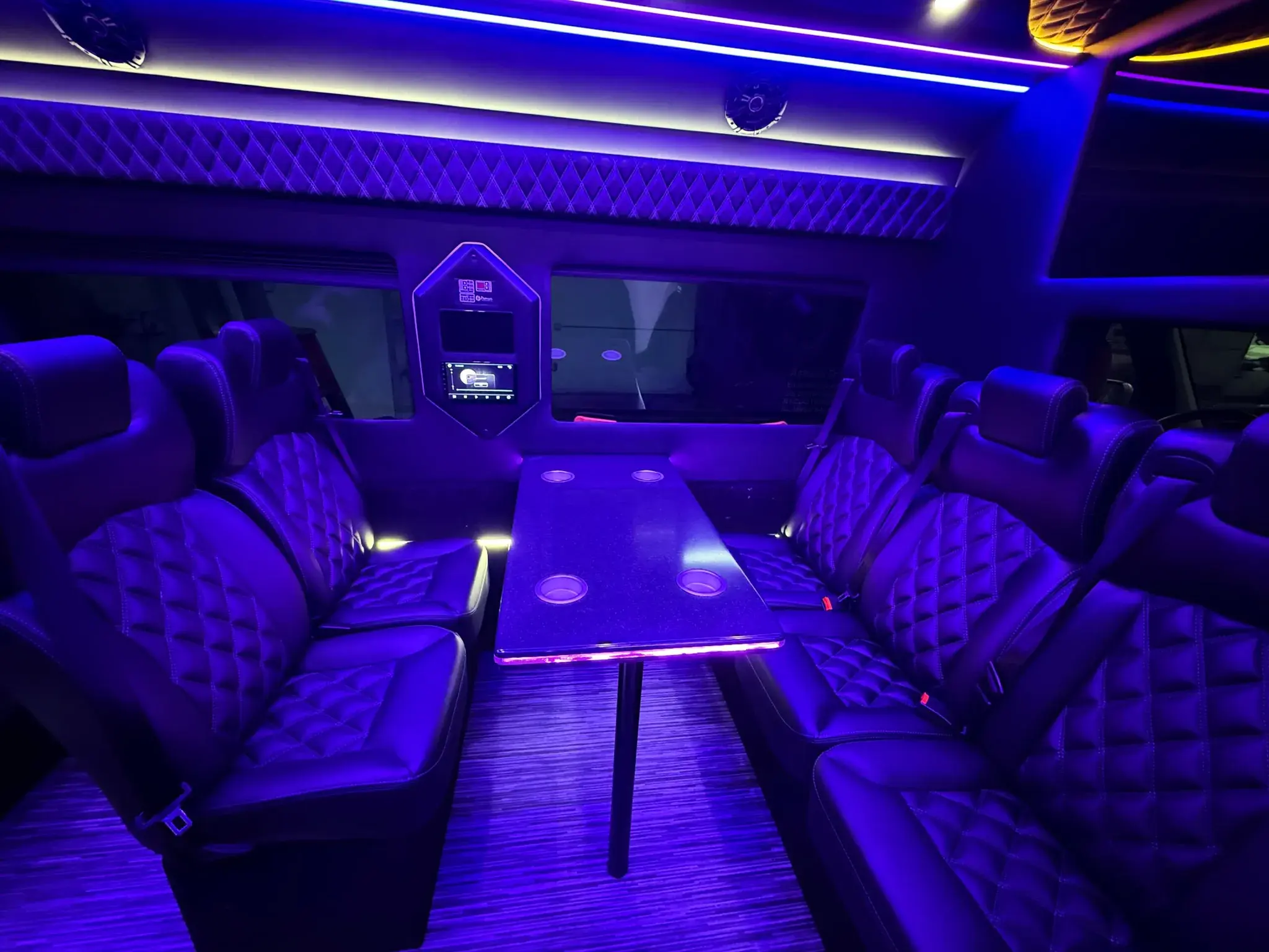Conference layout inside the Luxury Sprinter, with face-to-face quilted seating around a fixed table and ambient lighting along the roofline